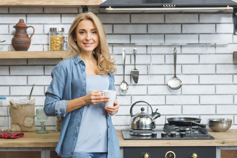 smiling blonde young woman standing near the gas stove holding white coffee cup